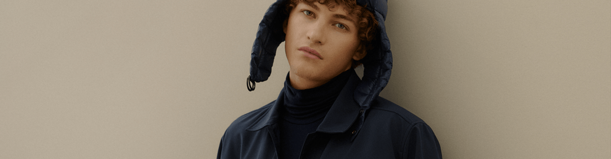 A model wearing a dark navy blue hooded jacket with ear flaps over a black turtleneck, set against a plain beige background.